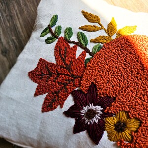 K&ouml;nnte beinhalten: Ein wei&szlig;es Kissen mit gestickten Herbstbl&auml;ttern und Blumen in Orange-, Braun-, Gelb-, Gr&uuml;n- und Burgunder-T&ouml;nen.