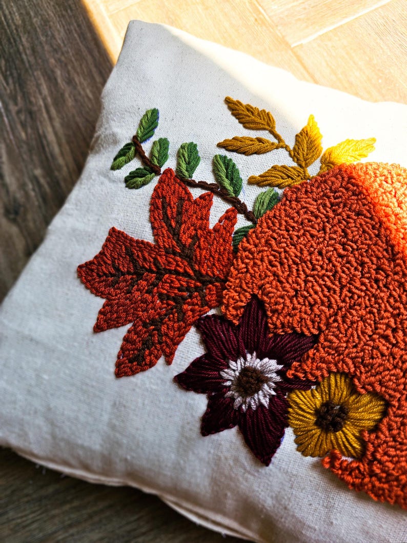 K&ouml;nnte beinhalten: Ein wei&szlig;es Kissen mit gestickten Herbstbl&auml;ttern und Blumen in Orange-, Braun-, Gelb-, Gr&uuml;n- und Burgunder-T&ouml;nen.