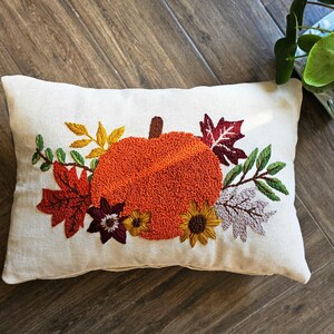 K&ouml;nnte beinhalten: Ein wei&szlig;es Kissen mit einer gestickten K&uuml;rbis, Bl&auml;ttern und Blumen in Orange-, Rot-, Gr&uuml;n- und Braunt&ouml;nen. Das Kissen ist perfekt f&uuml;r die Herbstdekoration.