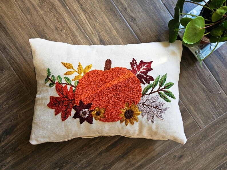 K&ouml;nnte beinhalten: Ein wei&szlig;es Kissen mit einer gestickten K&uuml;rbis, Bl&auml;ttern und Blumen in Orange-, Rot-, Gr&uuml;n- und Braunt&ouml;nen. Das Kissen ist perfekt f&uuml;r die Herbstdekoration.