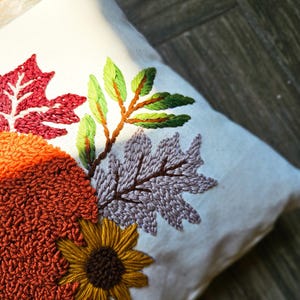 K&ouml;nnte beinhalten: Ein wei&szlig;es Kissen mit gestickten Herbstbl&auml;ttern in Rot-, Gr&uuml;n-, Lila-, Orange- und Braunt&ouml;nen. Das Kissen zeigt eine Sonnenblume mit brauner Mitte und gelben Bl&uuml;tenbl&auml;ttern.