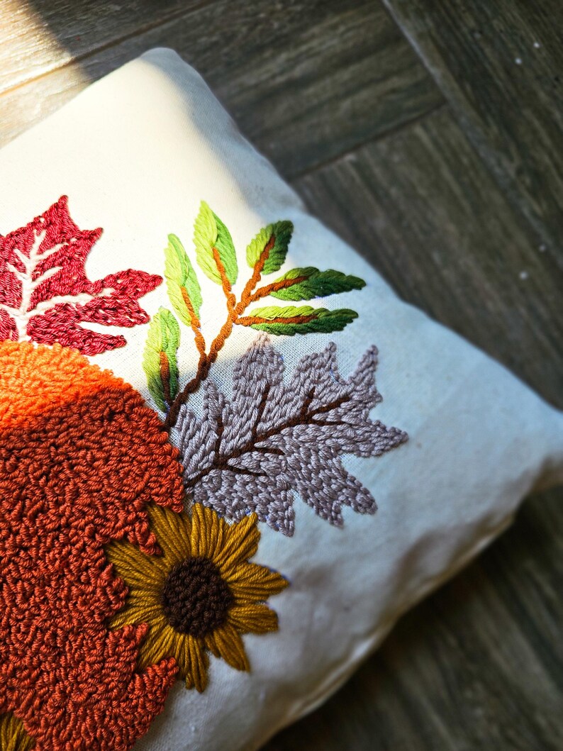 K&ouml;nnte beinhalten: Ein wei&szlig;es Kissen mit gestickten Herbstbl&auml;ttern in Rot-, Gr&uuml;n-, Lila-, Orange- und Braunt&ouml;nen. Das Kissen zeigt eine Sonnenblume mit brauner Mitte und gelben Bl&uuml;tenbl&auml;ttern.
