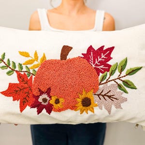 K&ouml;nnte beinhalten: Ein wei&szlig;es Kissen mit einem gestickten Design aus einem K&uuml;rbis, Bl&auml;ttern und Blumen in Orange-, Rot-, Gelb- und Gr&uuml;nt&ouml;nen.