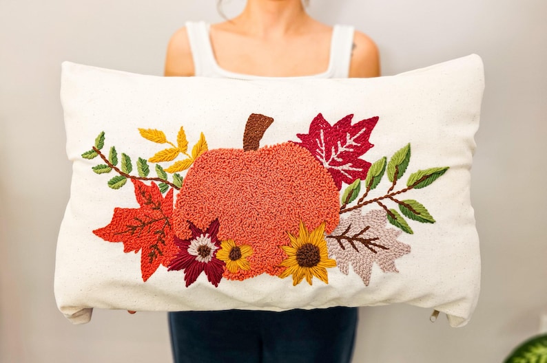 K&ouml;nnte beinhalten: Ein wei&szlig;es Kissen mit einem gestickten Design aus einem K&uuml;rbis, Bl&auml;ttern und Blumen in Orange-, Rot-, Gelb- und Gr&uuml;nt&ouml;nen.