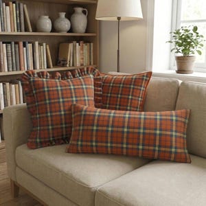 May include: Three decorative throw pillows featuring a rust orange, navy blue, and yellow plaid pattern. One square pillow has a ruffled edge, one is a standard square shape, and one is a rectangular lumbar pillow, all resting on a beige sofa.