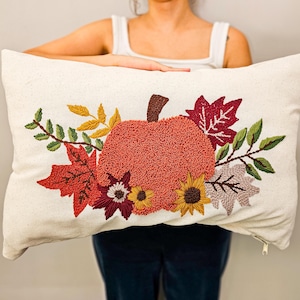 K&ouml;nnte beinhalten: Ein wei&szlig;es Kissen mit einem gestickten Design aus einem K&uuml;rbis, umgeben von Herbstbl&auml;ttern und Blumen. Der K&uuml;rbis ist orange und die Bl&auml;tter sind rot, orange und gelb.