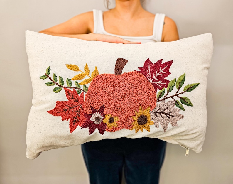 K&ouml;nnte beinhalten: Ein wei&szlig;es Kissen mit einem gestickten Design aus einem K&uuml;rbis, umgeben von Herbstbl&auml;ttern und Blumen. Der K&uuml;rbis ist orange und die Bl&auml;tter sind rot, orange und gelb.