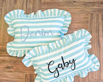 May include: Two blue and white striped pillow covers with ruffled edges. The pillow covers have the names "Denim" and "Gaby" embroidered on them.