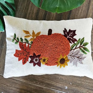 K&ouml;nnte beinhalten: Ein wei&szlig;es Kissen mit einer gestickten orangefarbenen K&uuml;rbis und Herbstbl&auml;ttern in Rot-, Orange-, Gelb- und Gr&uuml;nt&ouml;nen.