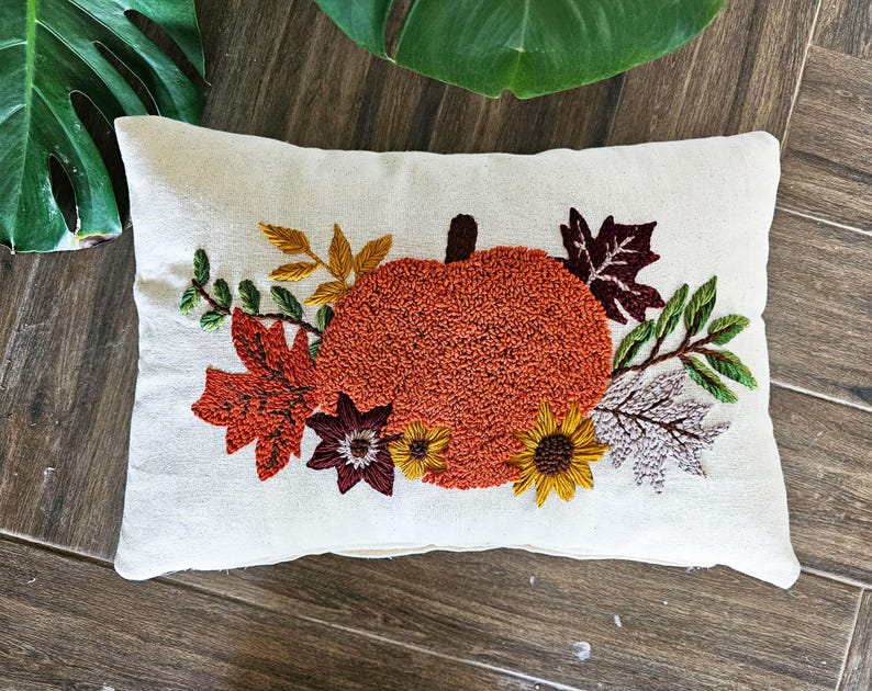 K&ouml;nnte beinhalten: Ein wei&szlig;es Kissen mit einer gestickten orangefarbenen K&uuml;rbis und Herbstbl&auml;ttern in Rot-, Orange-, Gelb- und Gr&uuml;nt&ouml;nen.
