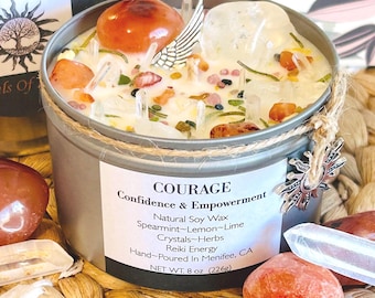 May include: A silver candle tin with a white candle inside. The candle is topped with crystals, herbs, and a silver angel wing charm. The label reads "COURAGE Confidence & Empowerment Natural Soy Wax Spearmint-Lemon-Lime Crystals-Herbs Reiki Energy Hand-Poured In Menifee, CA NET WT. 8 oz (226g)"