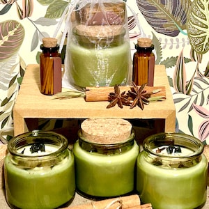 May include: Three green candles in glass jars with cork lids, accompanied by two small amber glass bottles filled with matches. A white organza bag holds a fourth green candle. Cinnamon sticks and star anise are arranged as decorative accents.