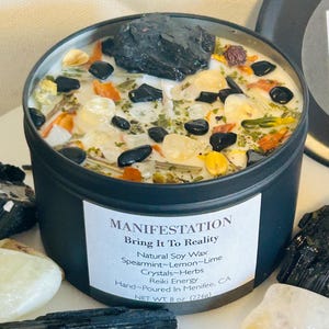 May include: Black candle tin with a white label that reads "MANIFESTATION Bring It To Reality Natural Soy Wax Spearmint-Lemon-Lime Crystals-Herbs Reiki Energy Hand-Poured In Menifee, CA NET WT. 8 oz (226g)". The candle is filled with white wax, black stones, clear crystals, and dried herbs.
