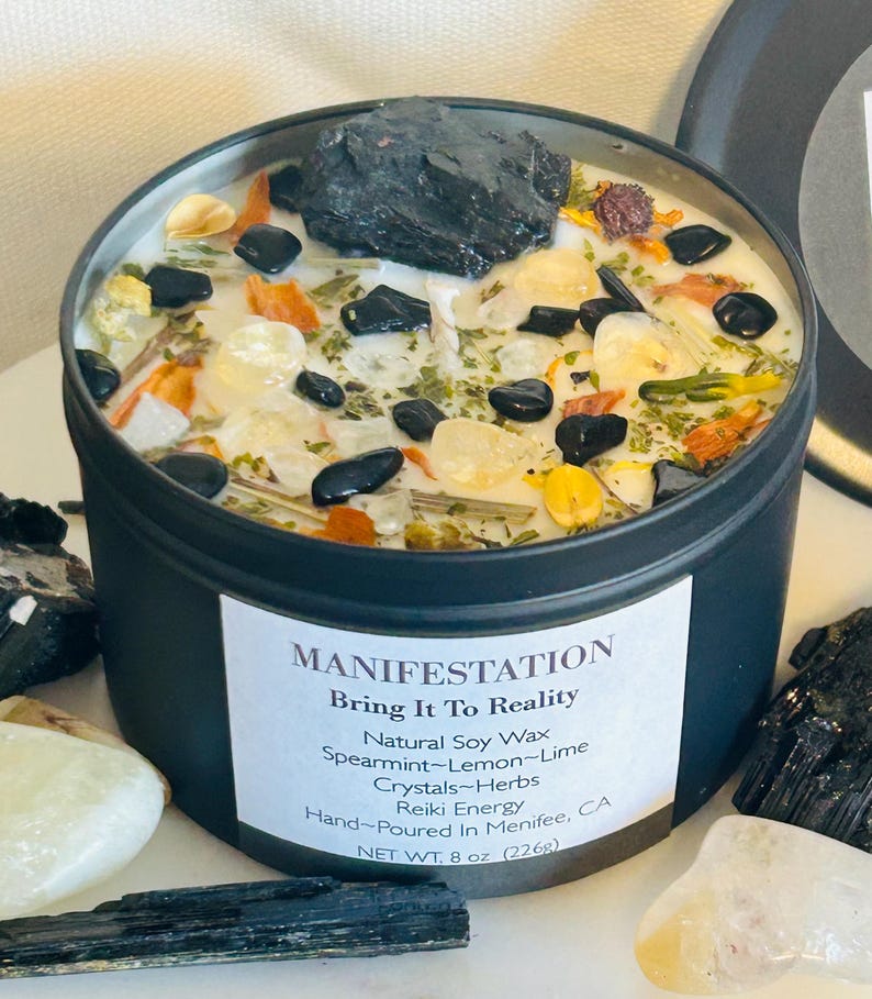 May include: Black candle tin with a white label that reads "MANIFESTATION Bring It To Reality Natural Soy Wax Spearmint-Lemon-Lime Crystals-Herbs Reiki Energy Hand-Poured In Menifee, CA NET WT. 8 oz (226g)". The candle is filled with white wax, black stones, clear crystals, and dried herbs.