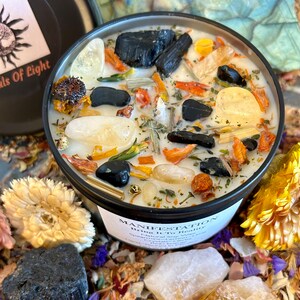 May include: A black candle jar with the words "MANIFESTATION Bring It To Reality" printed on the label. The candle is filled with white wax and contains various crystals, herbs, and flowers.