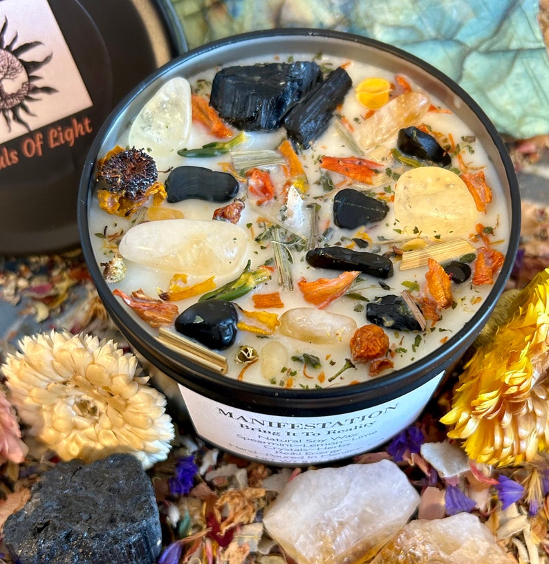 May include: A black candle jar with the words "MANIFESTATION Bring It To Reality" printed on the label. The candle is filled with white wax and contains various crystals, herbs, and flowers.