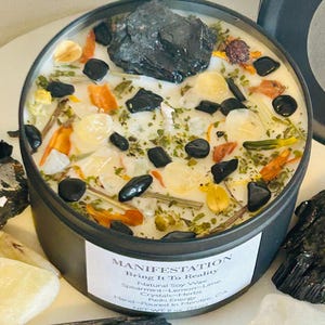 May include: A black candle jar with a white label that reads "MANIFESTATION Bring It To Reality" and a list of ingredients including "Natural Soy Wax", "Spearmint-Lemon-Lime", "Crystals-Herbs", "Reiki Energy", "Hand-Poured in Menifee CA", "NET WT 8 oz (226g)". The candle jar is filled with white wax and contains various herbs, crystals, and black stones.
