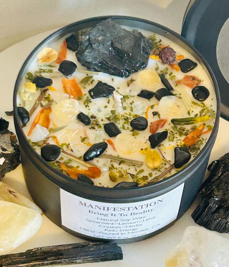 May include: A black candle jar with a white label that reads "MANIFESTATION Bring It To Reality" and a list of ingredients including "Natural Soy Wax", "Spearmint-Lemon-Lime", "Crystals-Herbs", "Reiki Energy", "Hand-Poured in Menifee CA", "NET WT 8 oz (226g)". The candle jar is filled with white wax and contains various herbs, crystals, and black stones.