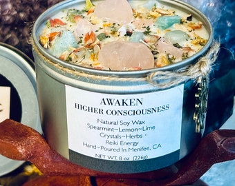 May include: A silver candle tin with a white label that reads "AWAKEN HIGHER CONSCIOUSNESS". The label lists the ingredients as "Natural Soy Wax, Spearmint-Lemon-Lime, Crystals-Herbs, Reiki Energy". The candle is hand-poured in Menifee, CA and weighs 8 oz (226g). The candle is topped with crystals and dried flowers.