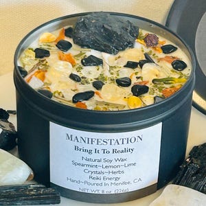 May include: Black candle tin with a white label that reads "MANIFESTATION Bring It To Reality Natural Soy Wax Spearmint-Lemon-Lime Crystals-Herbs Reiki Energy Hand-Poured In Menifee, CA NET WT. 8 oz (226g)". The candle is filled with white wax and has various crystals and herbs embedded in it.