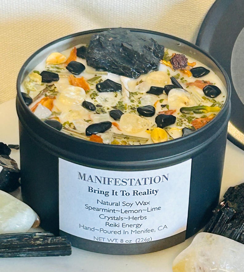May include: Black candle tin with a white label that reads "MANIFESTATION Bring It To Reality Natural Soy Wax Spearmint-Lemon-Lime Crystals-Herbs Reiki Energy Hand-Poured In Menifee, CA NET WT. 8 oz (226g)". The candle is filled with white wax and has various crystals and herbs embedded in it.