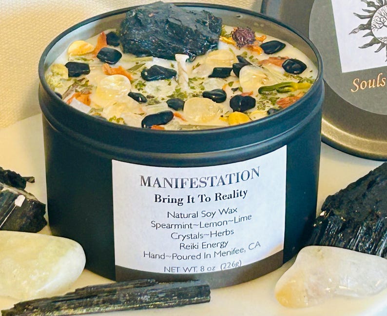 May include: Black metal candle tin with a white label that reads "MANIFESTATION Bring It To Reality Natural Soy Wax Spearmint-Lemon-Lime Crystals-Herbs Reiki Energy Hand-Poured In Menifee, CA NET WT. 8 oz (226g)". The candle is filled with white wax and includes black stones, green herbs, and small white crystals.
