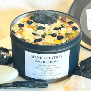 May include: A black candle tin with a white label that reads "MANIFESTATION Bring It To Reality Natural Soy Wax Spearmint-Lemon-Lime Crystals-Herbs Reiki Energy Hand-Poured In Menifee, CA NET WT. 8 oz (226g)". The candle is topped with a variety of crystals and herbs.