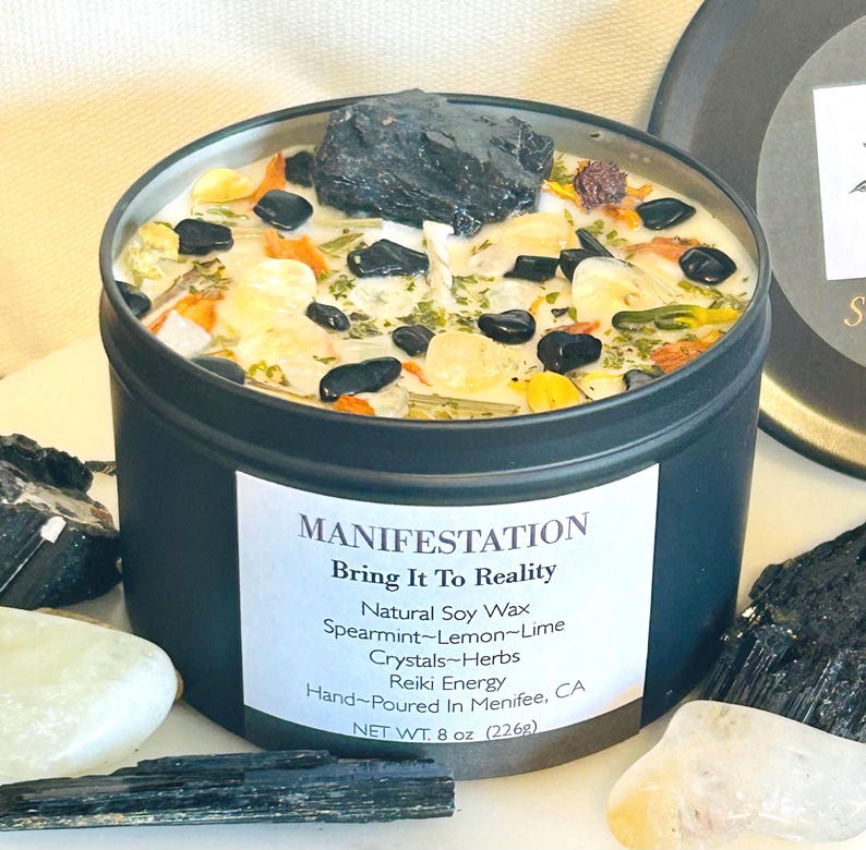 May include: A black candle tin with a white label that reads "MANIFESTATION Bring It To Reality Natural Soy Wax Spearmint-Lemon-Lime Crystals-Herbs Reiki Energy Hand-Poured In Menifee, CA NET WT. 8 oz (226g)". The candle is topped with a variety of crystals and herbs.