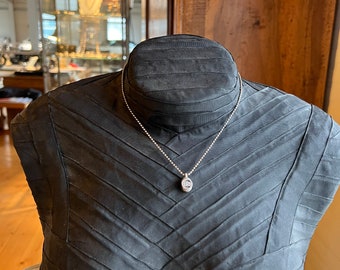 May include: A silver chain necklace with a small oval pendant featuring a clear gemstone. The necklace is displayed on a black mannequin.
