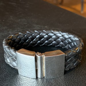Op de afbeelding: Een zwarte leren armband met een zilveren sluiting. De armband is gevlochten en heeft een eenvoudig ontwerp.