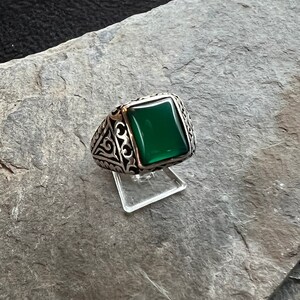 Könnte beinhalten: Ein silberner Ring mit einem verzierten Design und einem quadratischen grünen Edelstein.