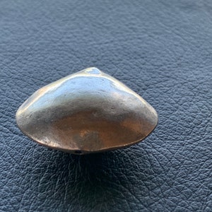 May include: A small, metallic clam shell. The shell is a muted gold color with a smooth, polished surface. The clam shell is resting on a dark, textured surface.
