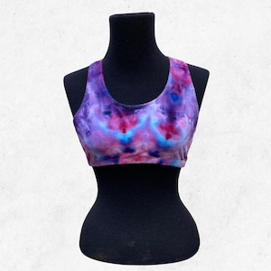 May include: A tie-dye sports bra in shades of purple, pink, and blue. The bra is displayed on a black mannequin. The fabric has a swirling, watercolor-like pattern.