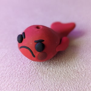 May include: A red clay figurine of a fish with a grumpy expression. The fish has black eyes, a frown, and a red blush on its cheek.