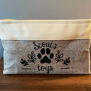 May include: A gray and white fabric storage bin with black text that reads "Scout's Toys" and a paw print.