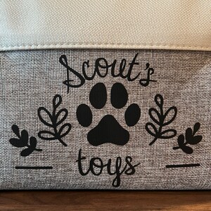 May include: A gray fabric storage bin with black text that reads "Scout's toys" and a black paw print. The bin has a white fabric top.