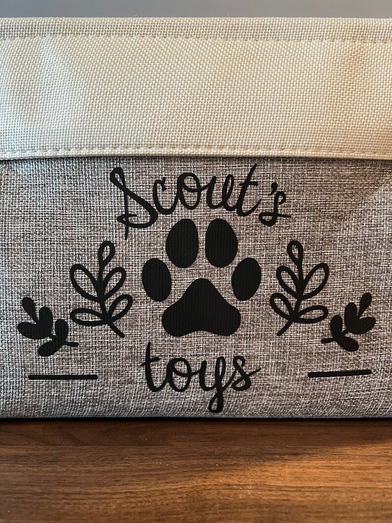 May include: A gray fabric storage bin with black text that reads "Scout's toys" and a black paw print. The bin has a white fabric top.