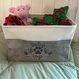 May include: A gray and white storage bin with the text "Bailey's toys" and a paw print design. The bin is filled with various dog toys, including a pink pig, a red and black ring, and a green and red rope toy.