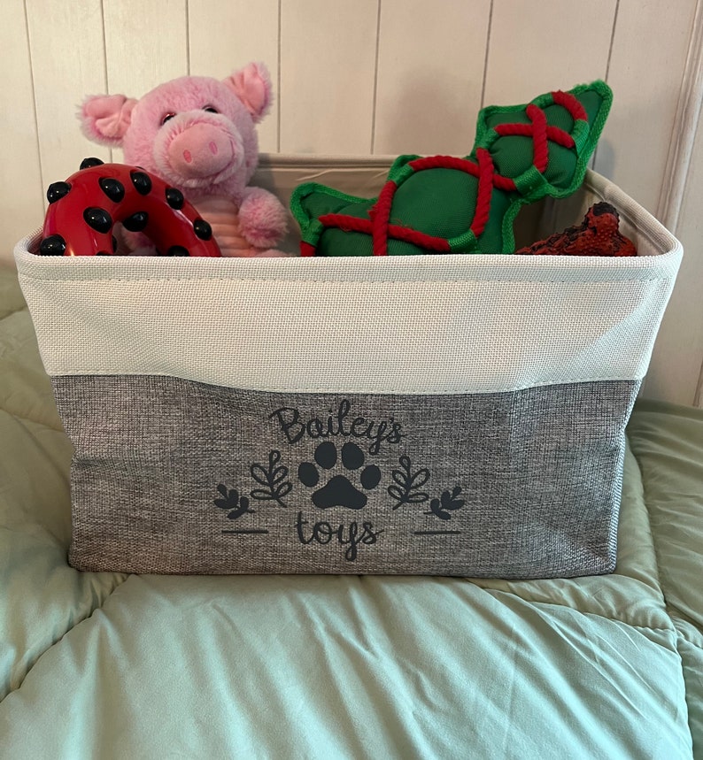 May include: A gray and white storage bin with the text "Bailey's toys" and a paw print design. The bin is filled with various dog toys, including a pink pig, a red and black ring, and a green and red rope toy.