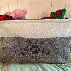 May include: A gray and white storage bin with the text "Bailey's toys" and a black paw print design. The bin is filled with various stuffed toys, including a pink pig and a red and green toy.