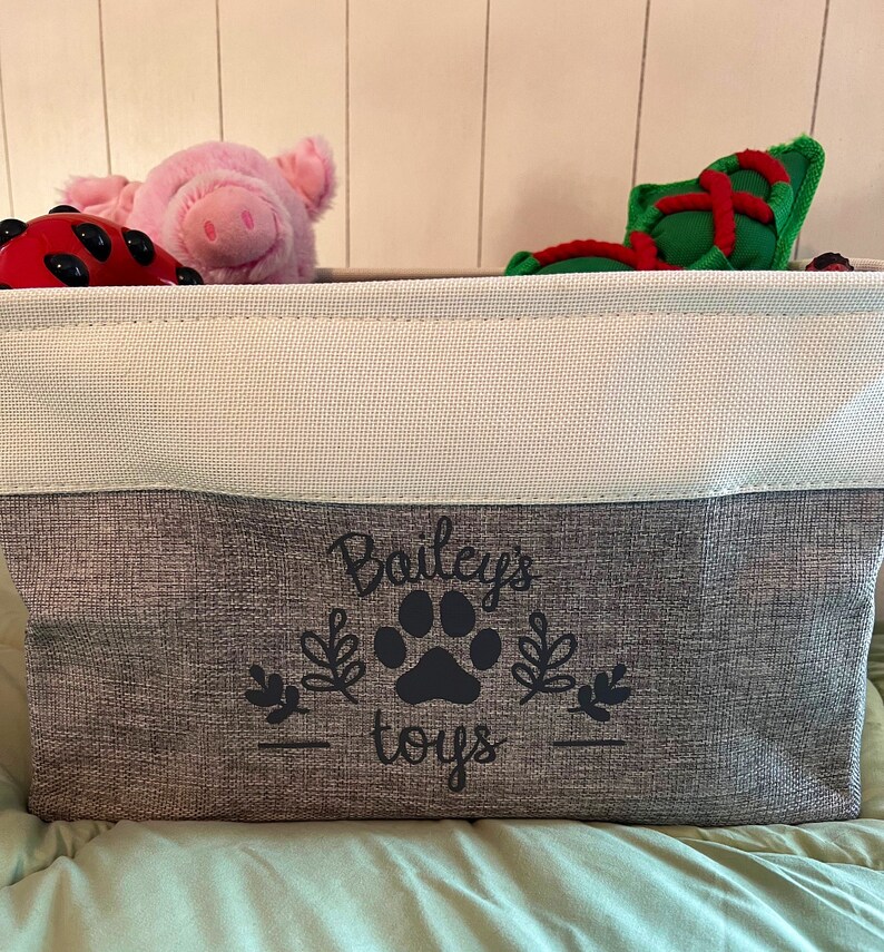 May include: A gray and white storage bin with the text "Bailey's toys" and a black paw print design. The bin is filled with various stuffed toys, including a pink pig and a red and green toy.