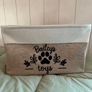 May include: A beige and white storage bin with a black paw print and the text "Bailey's toys" on the front.