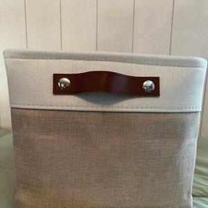 May include: A light gray and brown fabric storage bin with a brown leather handle and silver metal accents. The bin has a rectangular shape and is made of a woven fabric.