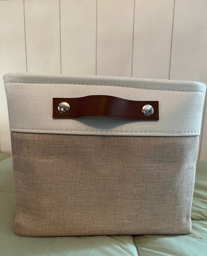 May include: A light gray and brown fabric storage bin with a brown leather handle and silver metal accents. The bin has a rectangular shape and is made of a woven fabric.