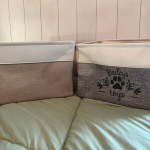 May include: Two fabric storage bins with white tops and gray bottoms. The bin on the right has the text "Bailey's toys" with a paw print in black.