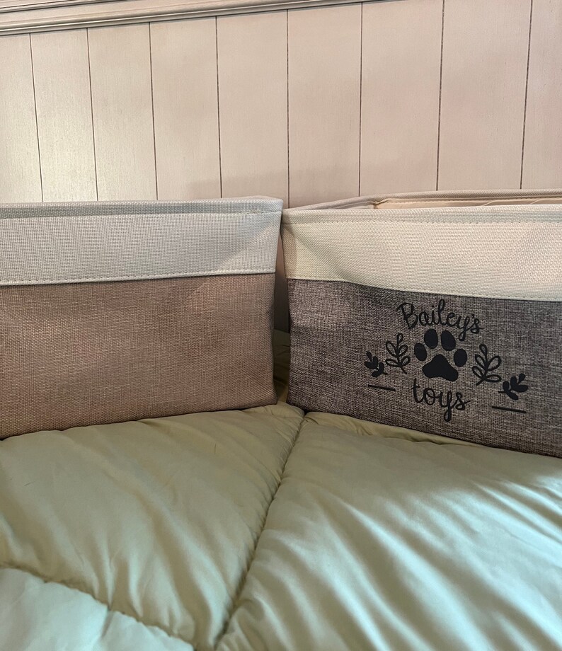 May include: Two fabric storage bins with white tops and gray bottoms. The bin on the right has the text "Bailey's toys" with a paw print in black.