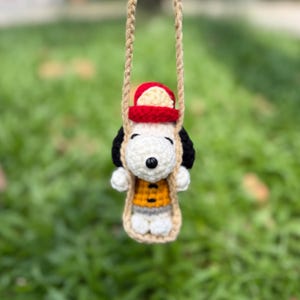 May include: A crocheted white dog figure with black ears, wearing a red hat and an orange vest with black buttons, sitting on a tan crocheted swing suspended by two braided tan ropes. The figure has a black nose and small white paws.