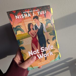 May include: A paperback book titled "Not Safe Work" by Nisha J. Tuli. The cover features a couple in a romantic setting with palm trees and a building in the background. The book has yellow sticky notes on the side.
