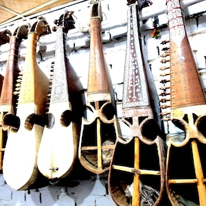 Puede incluir: Una fila de instrumentos musicales de cuerda de madera colgados verticalmente. Cada instrumento presenta un mástil largo, un cuerpo hueco con aberturas y patrones tallados intrincados. Los tonos de madera varían de claro a oscuro.