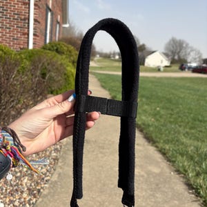 May include: Black shoulder strap with a soft, fuzzy pad and adjustable webbing. The strap has two silver-coloured swivel clips for attachment. The background shows a pavement, greenery, and a house.